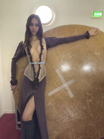 Model leaning against a large wooden disc wearing a plunging dress with ruffles and dark boots for Vivienne Westwood.