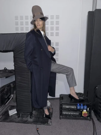 Model wearing a tall top hat and dark coat resting foot on equipment case in a backstage setting