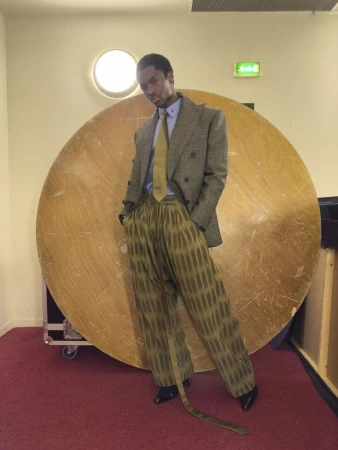 Black man standing in patterned trousers and tweed blazer against a large circular wooden prop on red carpet