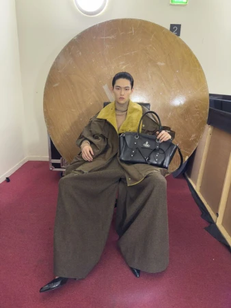 Model seated in front of a large circular wooden disc holding a black handbag for Vivienne Westwood