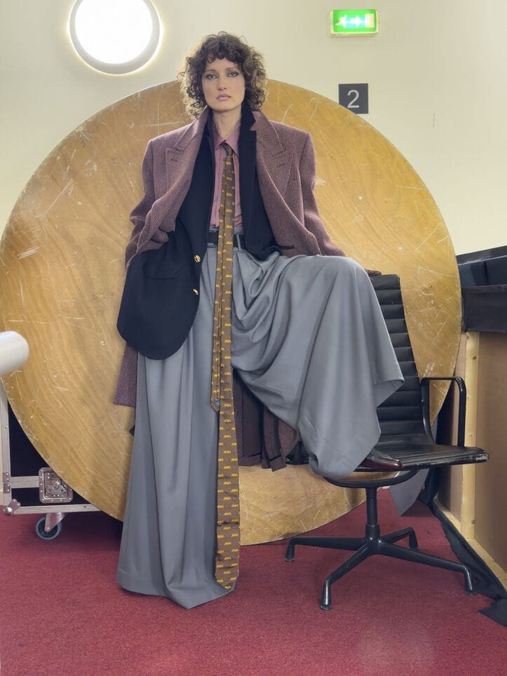 Model with curly hair wearing oversized layered tailoring and a patterned tie against a circular wooden backdrop
