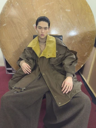 Model wearing an oversized olive green coat with a yellow collar seated against a large circular wooden disc.