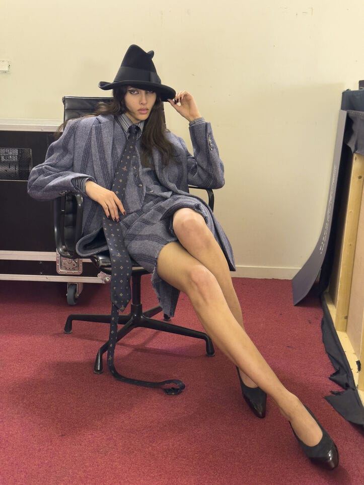 Model wearing a grey patterned suit and black witch hat seated on an office chair against a plain wall