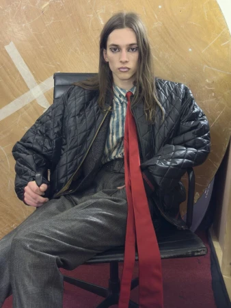 Model seated in an office chair wearing a black quilted bomber jacket, striped shirt, and a long red tie for Vivienne Westwood.