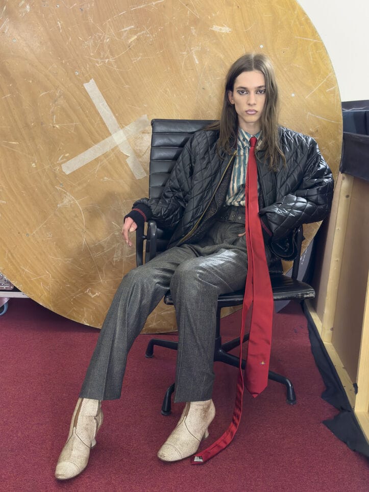 Model seated in an office chair wearing a black quilted jacket and long red tie against a large wooden circular backdrop