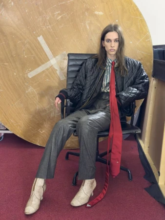 Model seated in an office chair wearing a black quilted jacket and long red tie against a large wooden circular backdrop