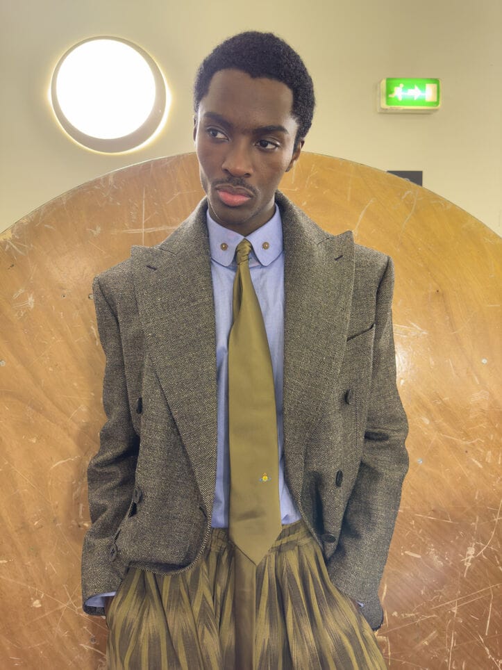 A black man wearing a grey tweed double-breasted jacket, light blue shirt, and olive tie in front of a wooden circular structure.