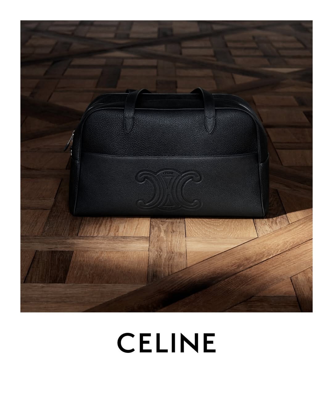 Black leather duffle bag with embossed logo resting on patterned wood parquet flooring