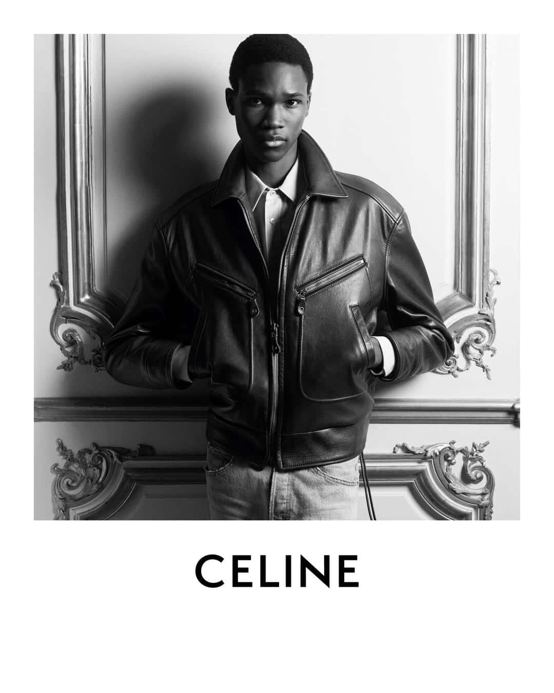 Black and white portrait of a male model wearing a leather jacket leaning against an ornate wall for Celine Homme Winter 2025.