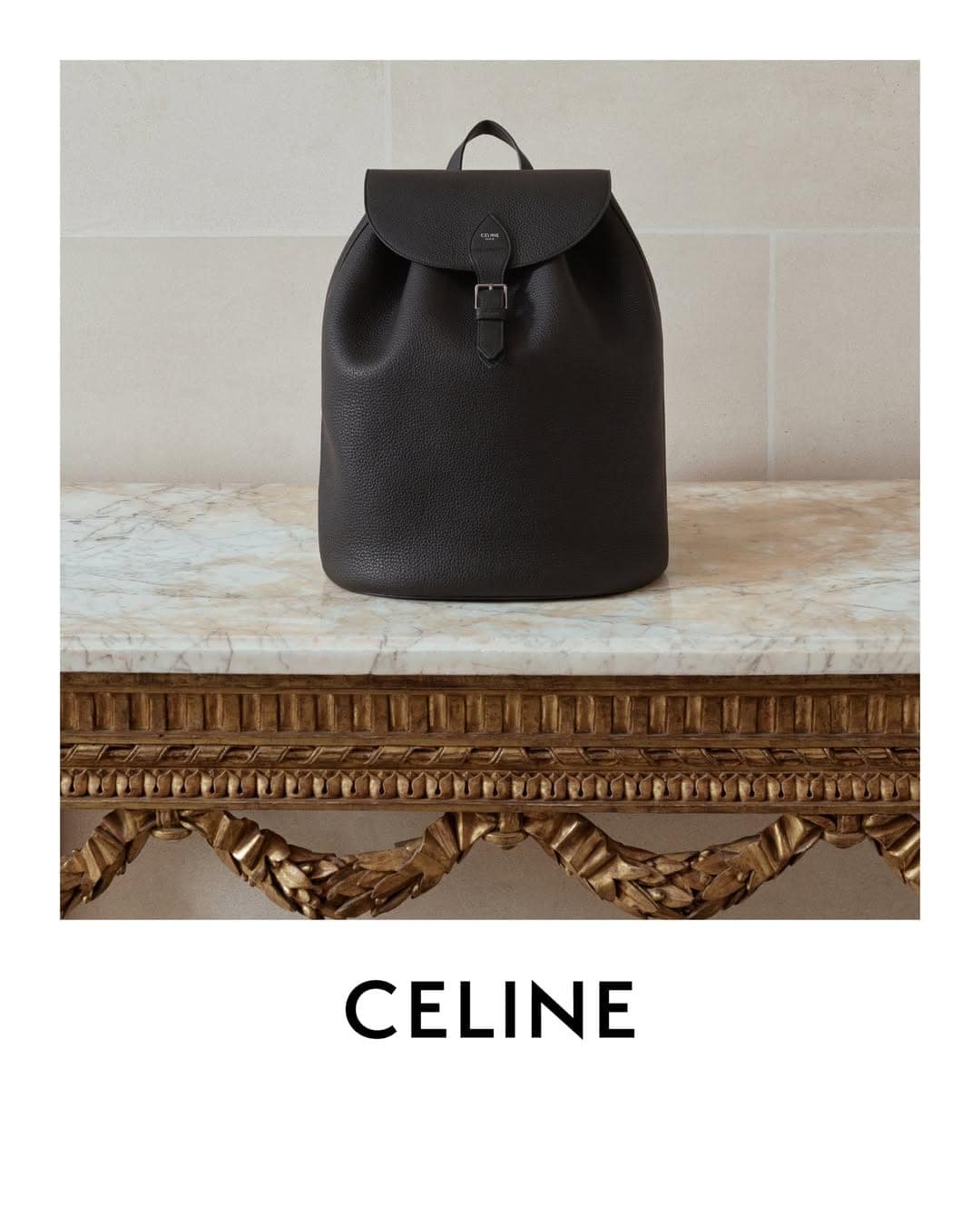 Dark brown leather backpack centered on an ornate gold console table against a neutral stone wall.