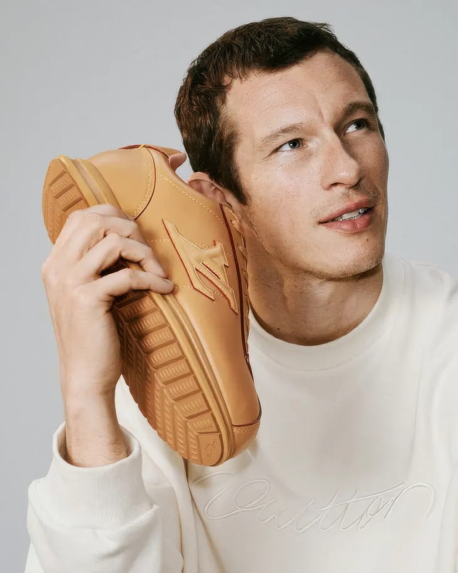 Close-up portrait of Callum Turner holding a tan leather sneaker to his ear like a phone, wearing a white sweatshirt, on grey background