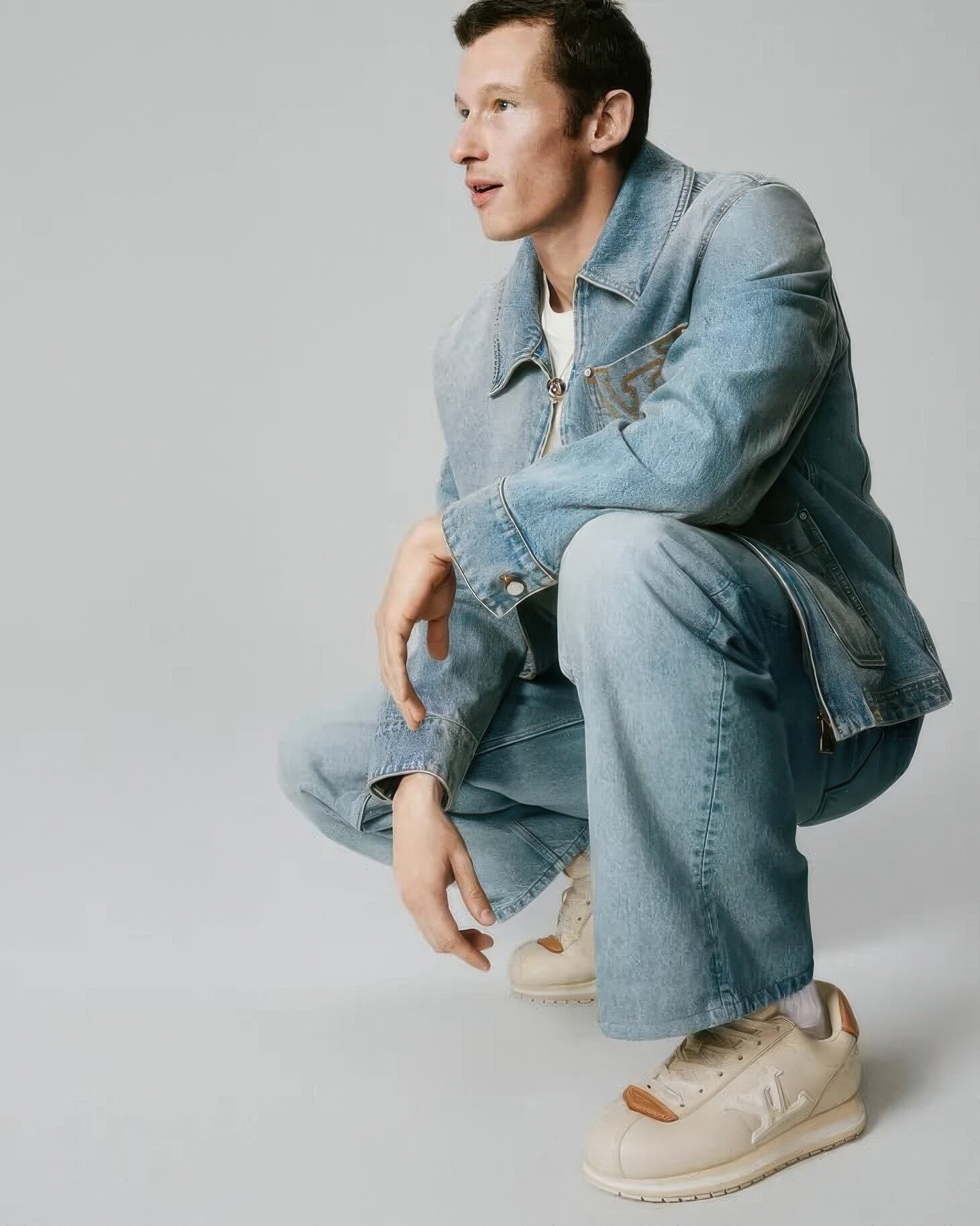 Model Callum Turner crouching in a denim jacket and light sneakers against a neutral grey studio background