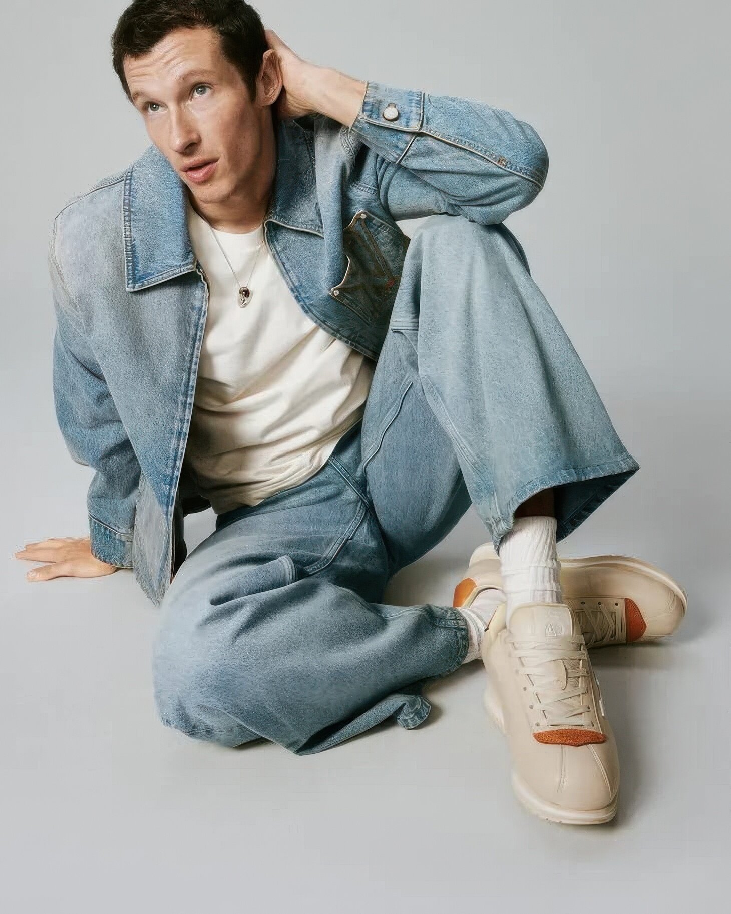 Model Callum Turner sitting on a grey floor wearing a denim jacket, white t-shirt, loose jeans, and Louis Vuitton buttersoft sneakers