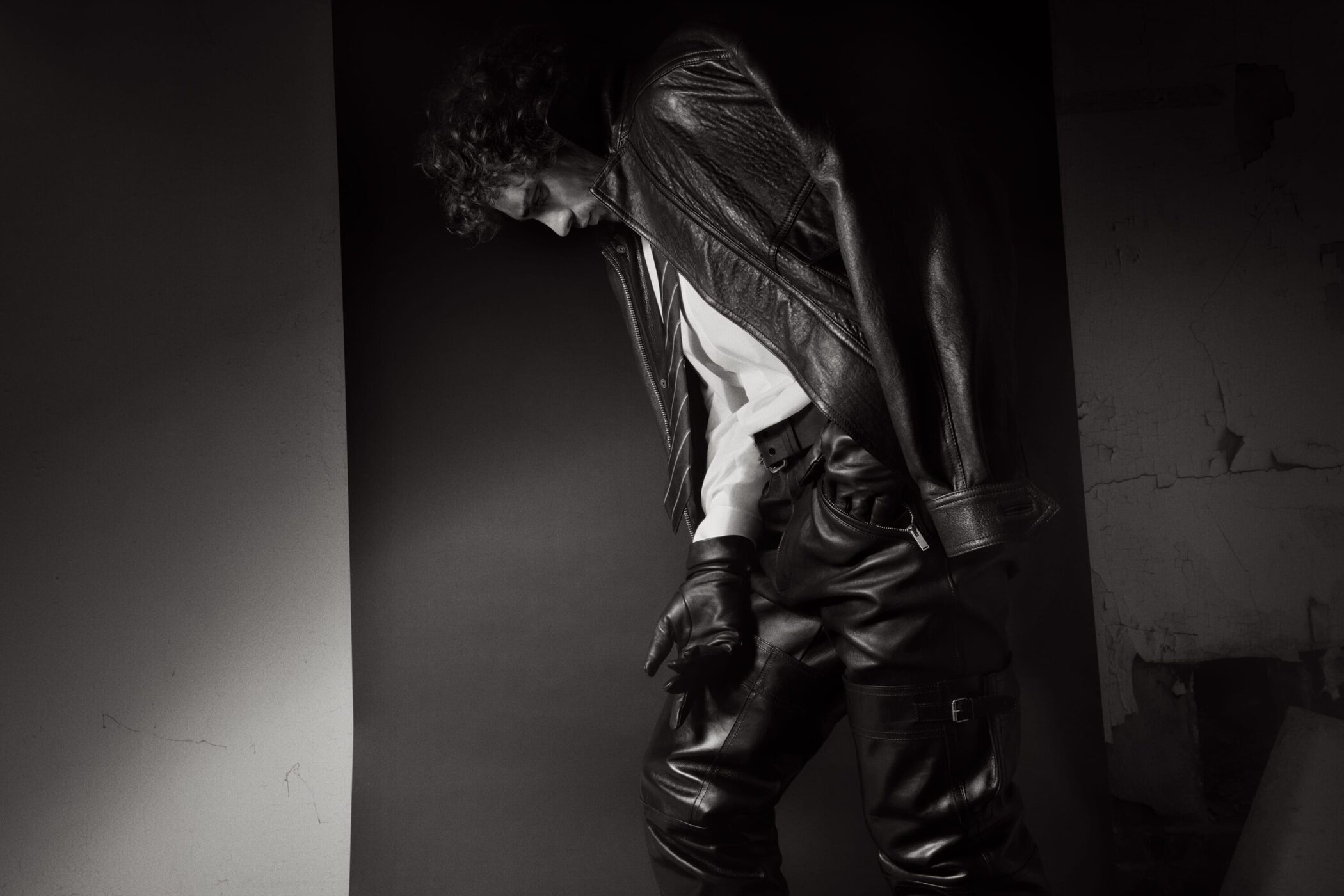Man wearing a leather jacket, white shirt, tie, and leather trousers against a dark backdrop in a black and white photograph.