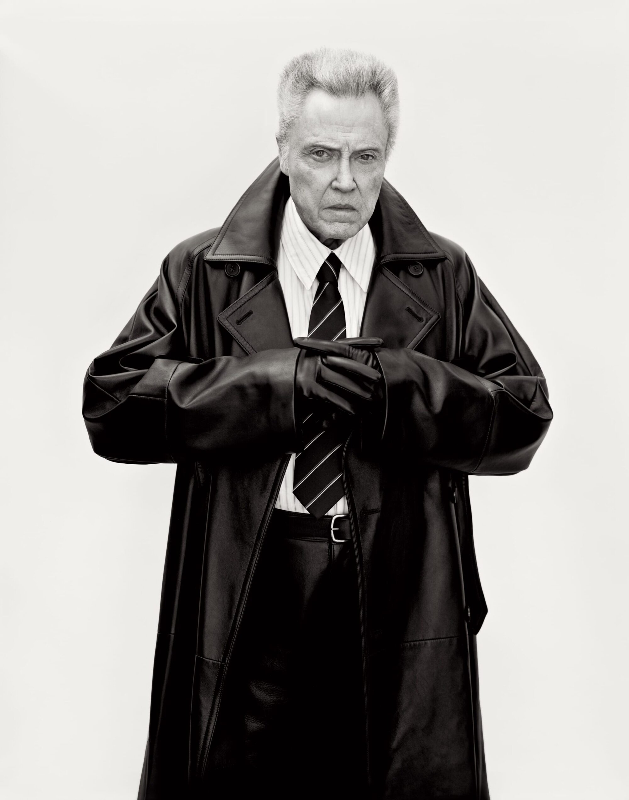 Christopher Walken in a black leather trench coat, striped shirt, and tie against a white background for Saint Laurent Winter 2025.