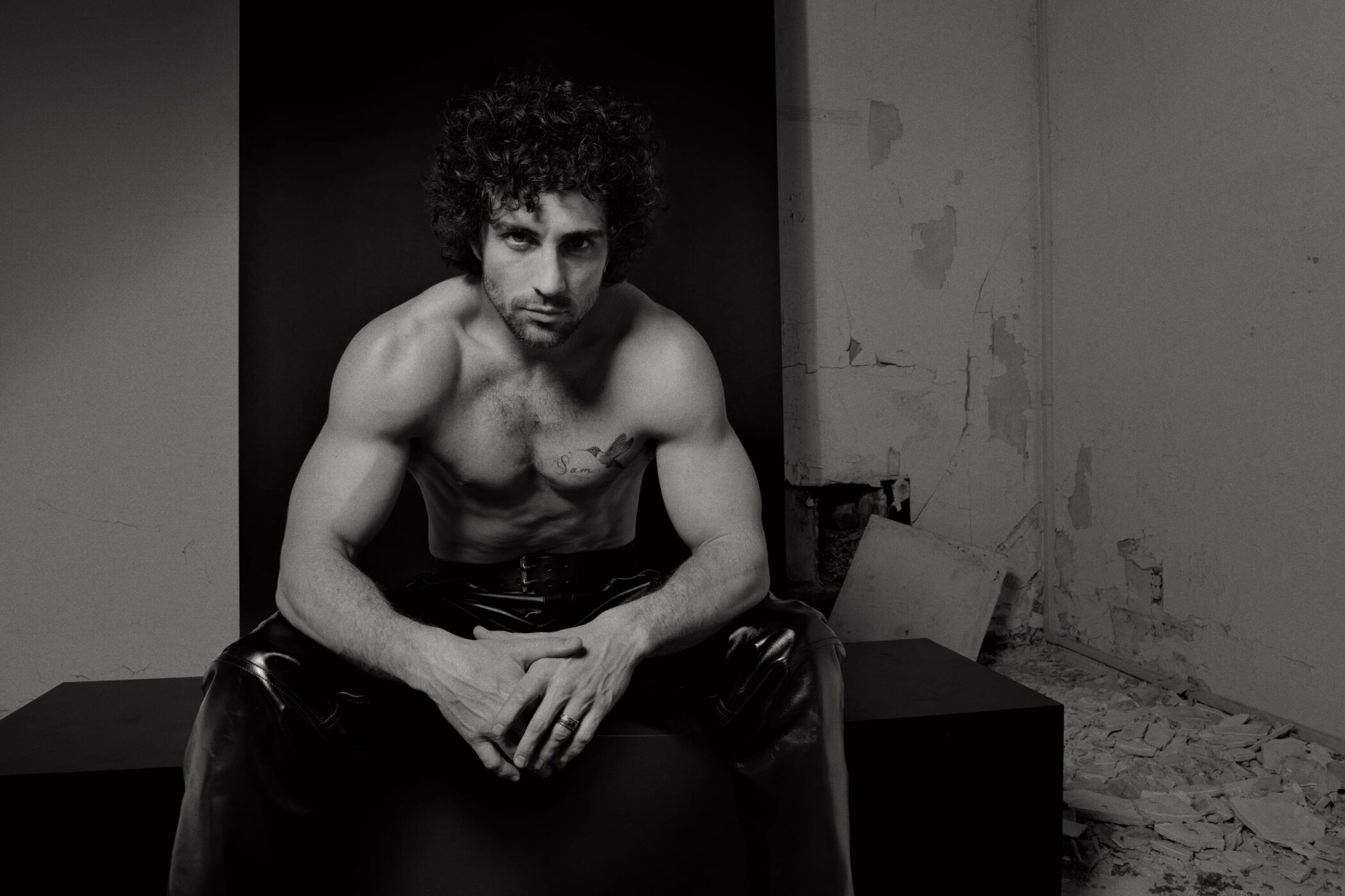 Monochrome portrait of Aaron Taylor-Johnson shirtless with dark trousers seated in an interior room