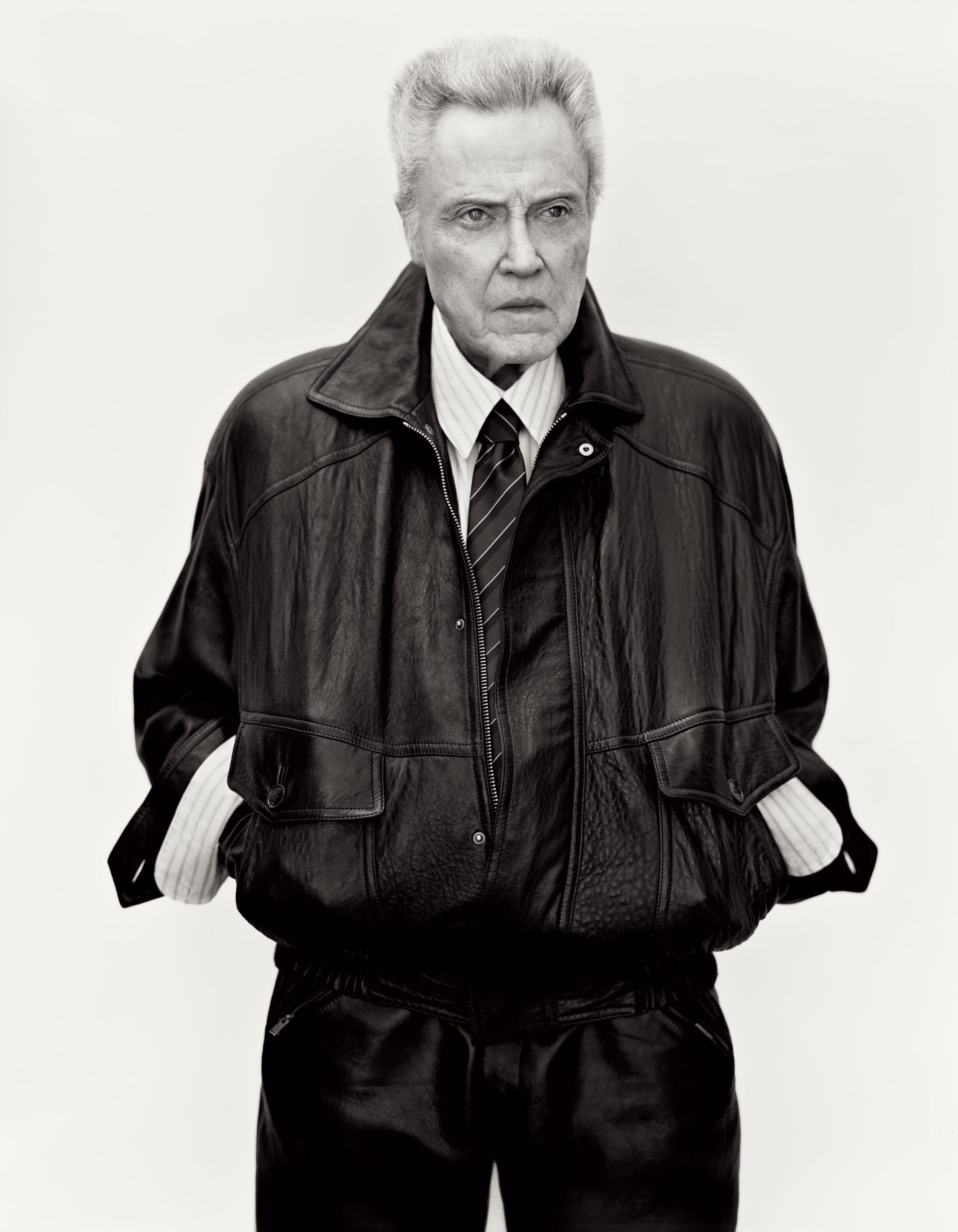 Christopher Walken wearing a leather bomber jacket, striped shirt, and tie against a seamless white background for Saint Laurent.