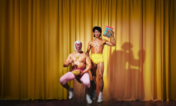 Two men posing against a gold curtain backdrop, one wearing a luchador mask and eating from a bowl with chopsticks