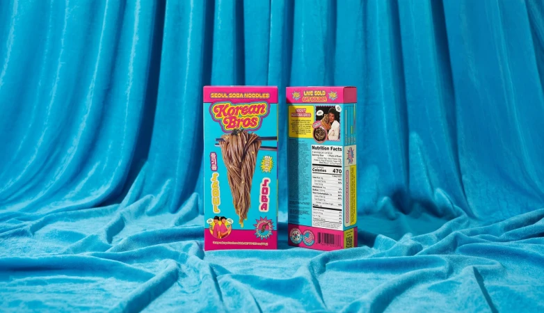 Two colorful product boxes for Korean Bros Soba noodles standing upright against a draped blue fabric backdrop.