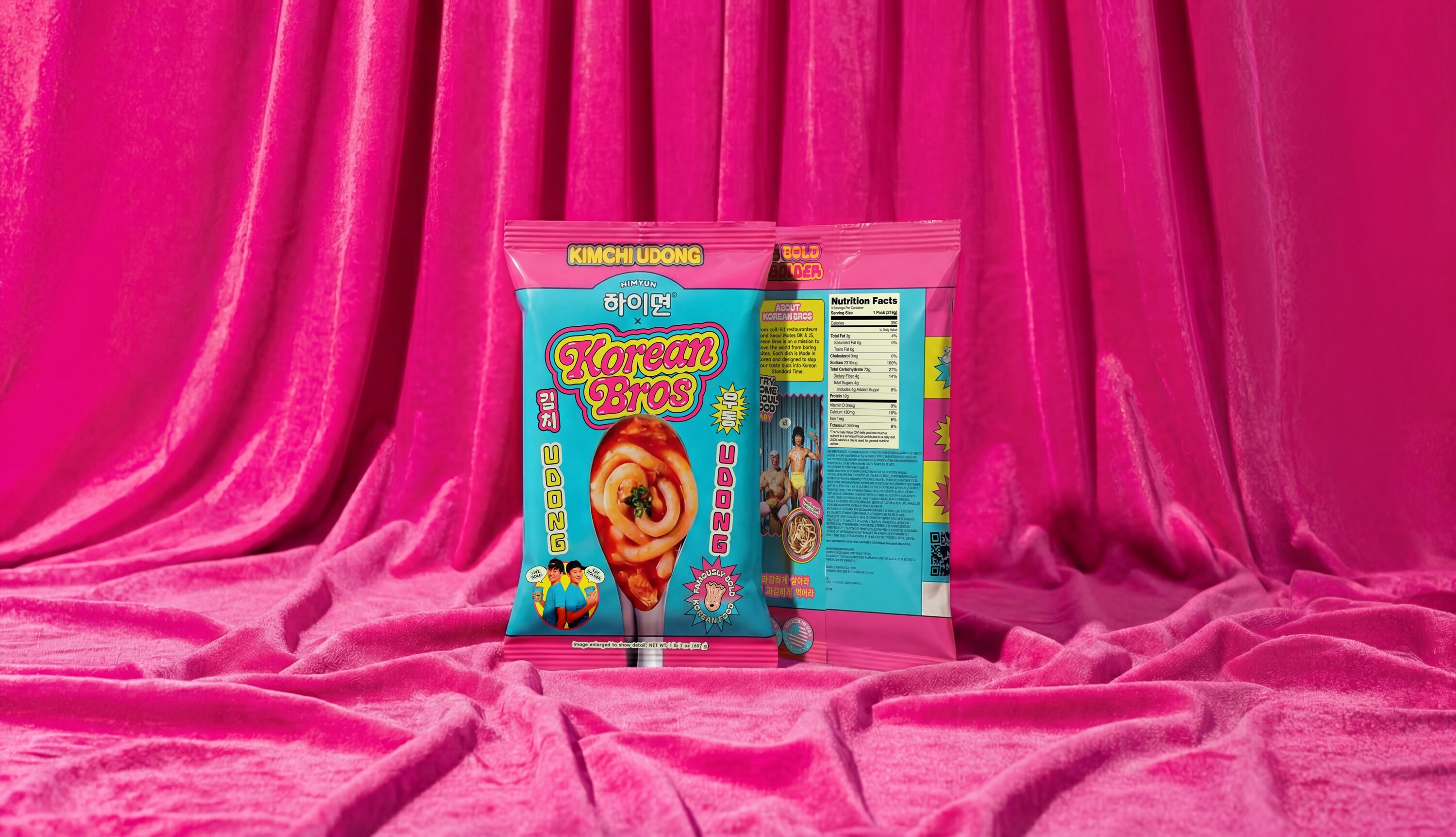 Two bright blue and pink product packages for Korean Bros Kimchi Udong displayed against a vibrant pink velvet fabric backdrop.