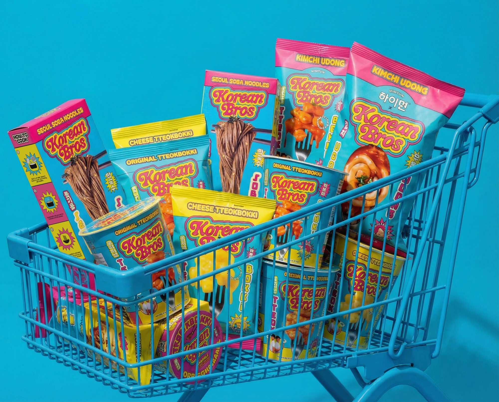 Assorted brightly colored Korean Bros noodle and tteokbokki packaging piled inside a light blue metal shopping cart against a blue backdrop.
