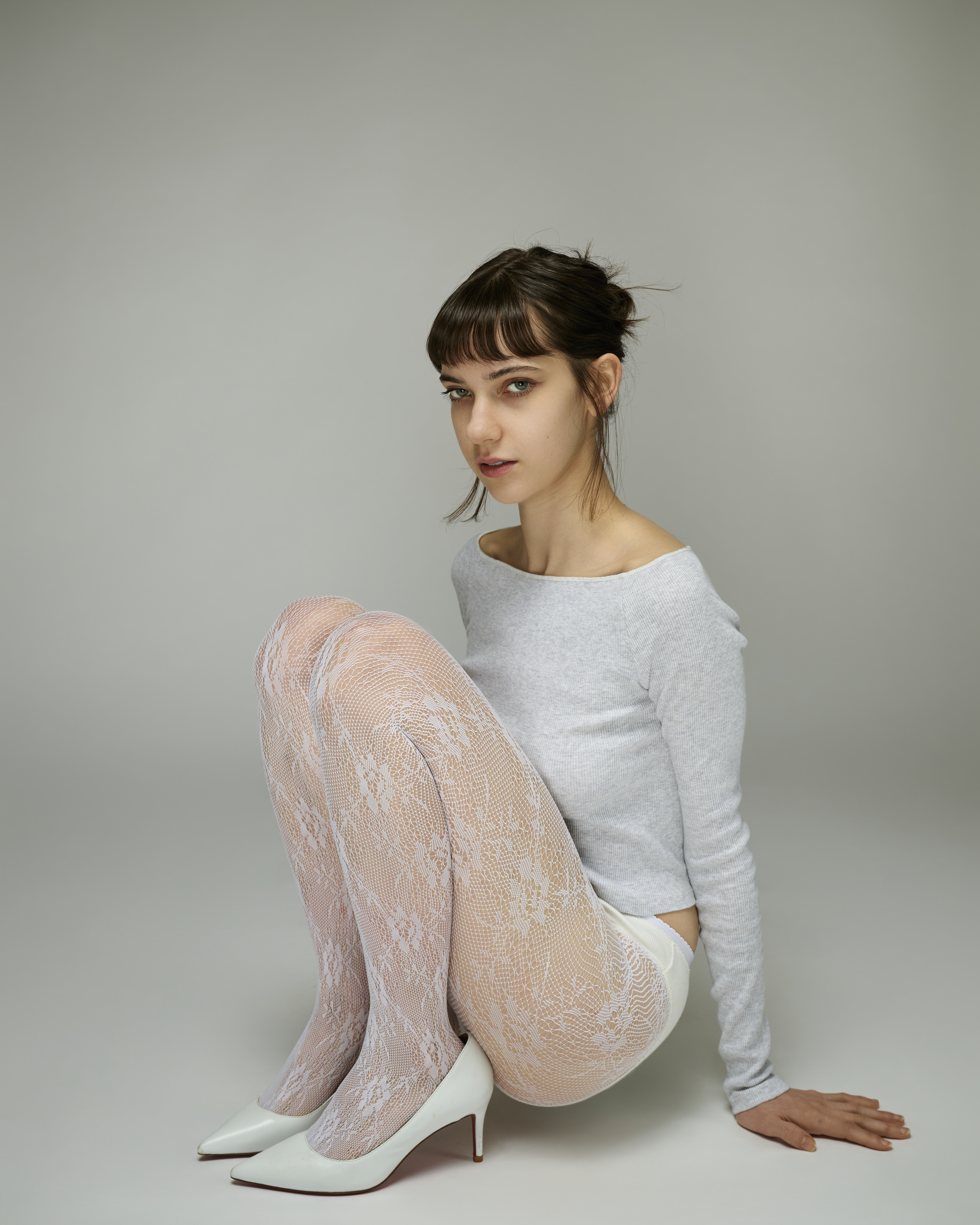 Model seated on white background wearing a light grey long-sleeve top, white lace tights, and white high heels.