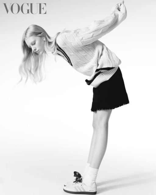Monochrome fashion shot of a blond woman in a sweater and skirt, bending over, showcasing athletic shoes in a bright white studio.