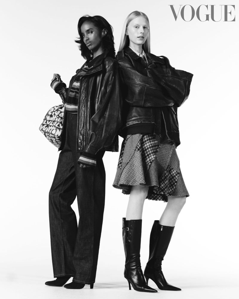 Two fashion models in a studio setting, wearing black leather jackets, skirts, jeans, and boots, posing back to back for Vogue magazine.