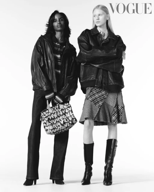 Two fashion models in black and white. The models are wearing leather jackets, with one carrying a graphic handbag in a white studio.