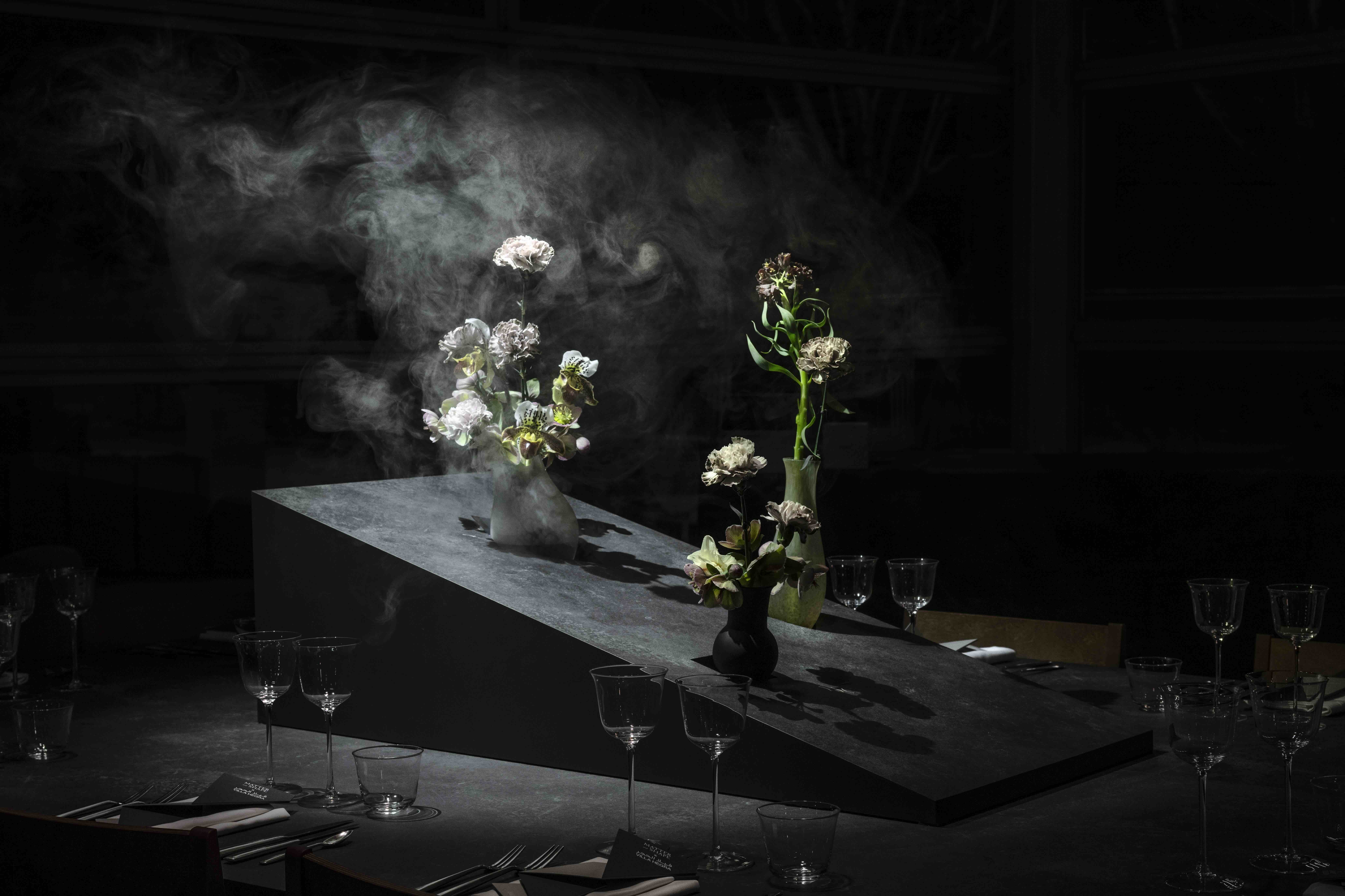 Dark still life photography of flower arrangements in vases on an angled dark slab with smoke drifting above, created by Matter and Shape.