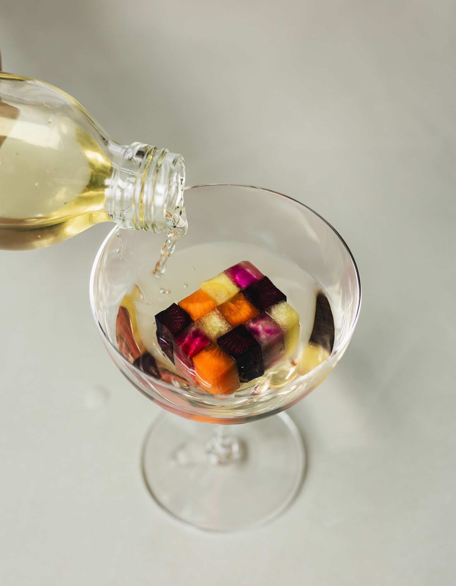 A clear glass being poured with liquid from a glass bottle, containing a colorful mosaic ice cube made of small vegetable squares.