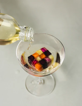 A clear glass being poured with liquid from a glass bottle, containing a colorful mosaic ice cube made of small vegetable squares.