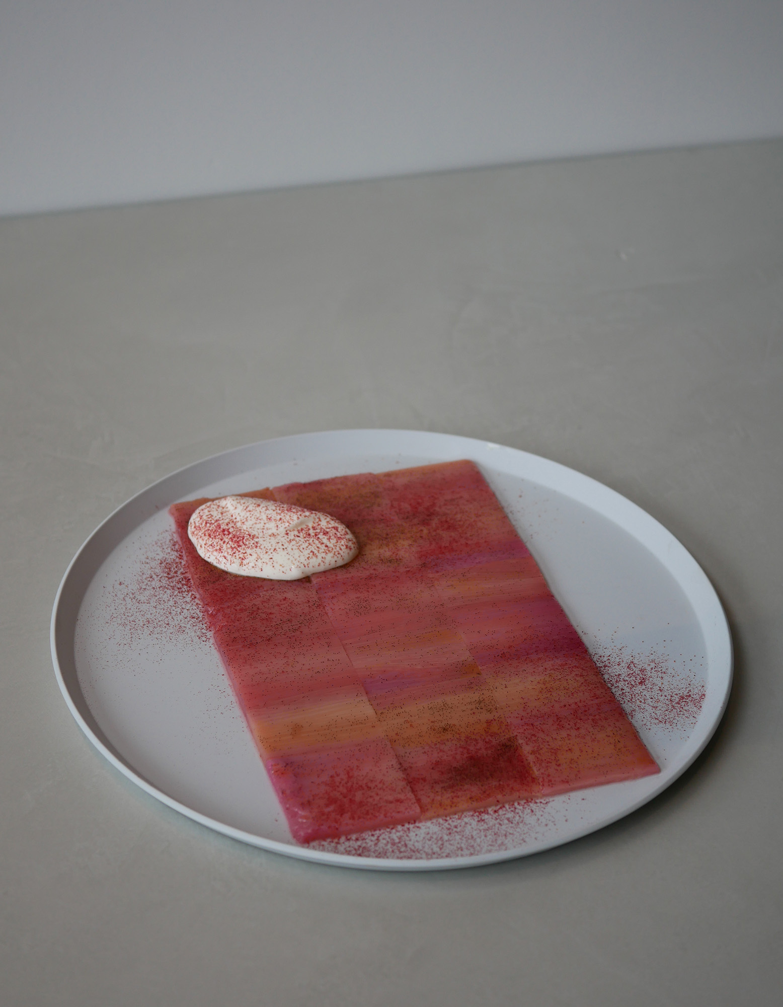 A white plate holding pink sliced food topped with white cream and red powder on a neutral grey surface.