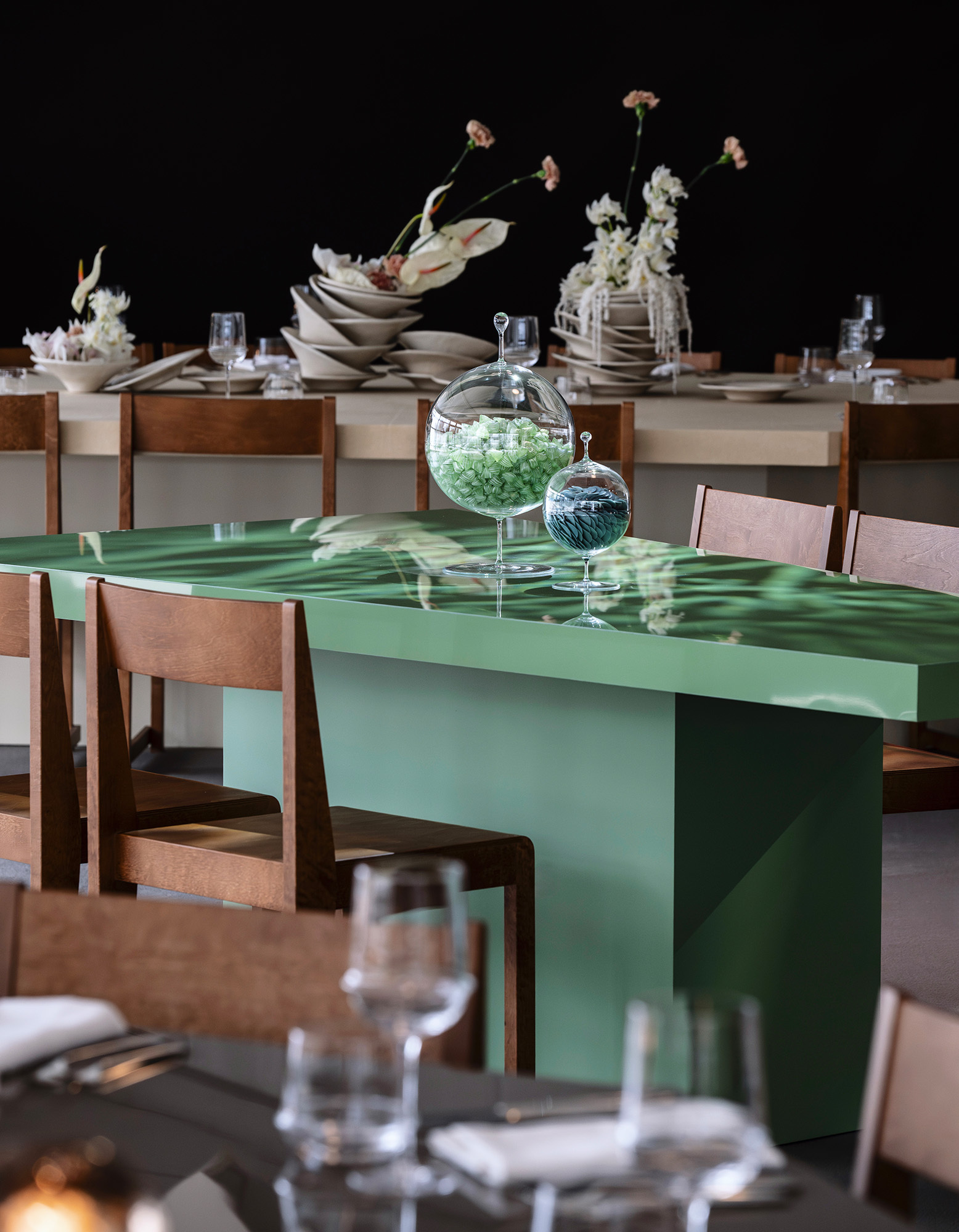 Green glossy dining table with glass cloches and wooden chairs set in an interior restaurant space by Matter and Shape.