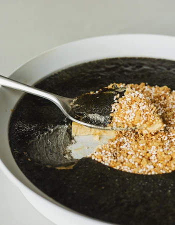 Silver spoon scooping a portion of dark textured food topped with grains from a large white bowl.
