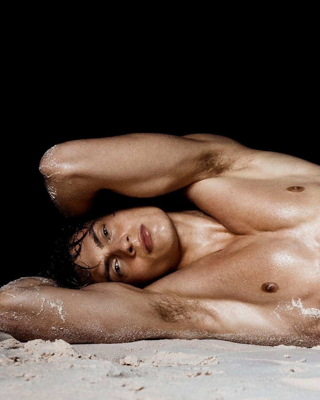 Close-up portrait of a shirtless man lying down with sand on his skin against a dark background for Calvin Klein.