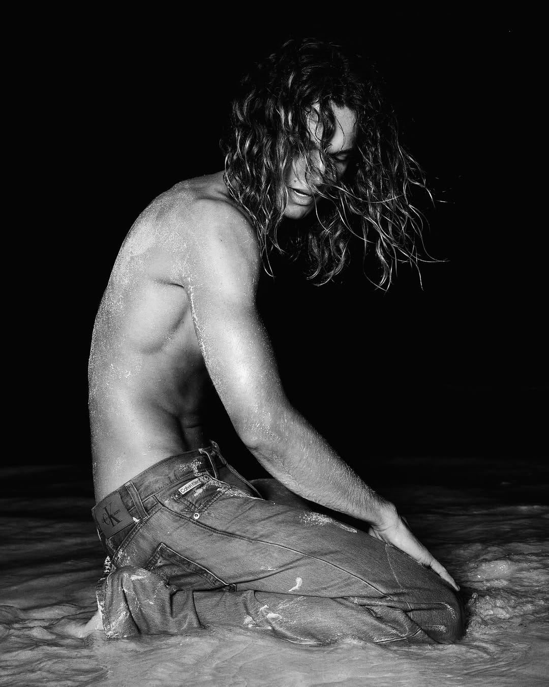 Monochrome image of a shirtless man with long curly hair kneeling in shallow water wearing denim jeans for Calvin Klein.
