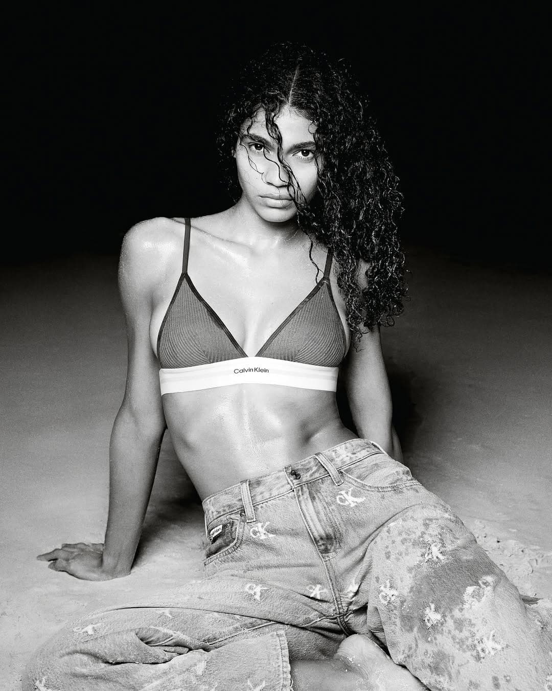 Model with dark curly hair wearing a Calvin Klein bralette and monogram denim jeans seated on a dark ground.