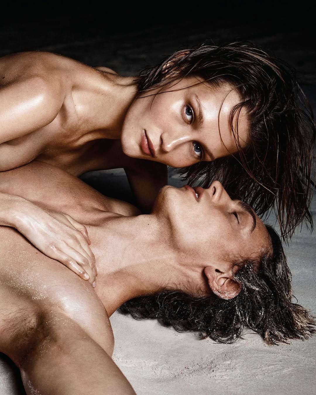 Close-up of a man and woman lying close together on pale sand with wet skin and hair, photographed by Stuart Winecoff.