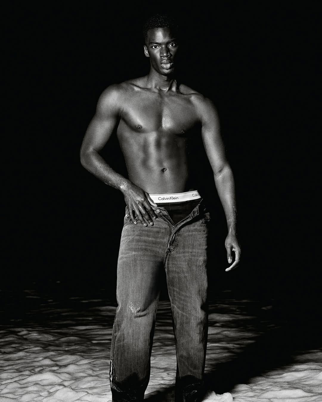 Shirtless black man wearing denim jeans standing against a dark background for Calvin Klein Summer 2025 Ad Campaign.