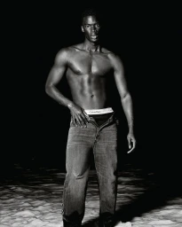 Shirtless black man wearing denim jeans standing against a dark background for Calvin Klein Summer 2025 Ad Campaign.