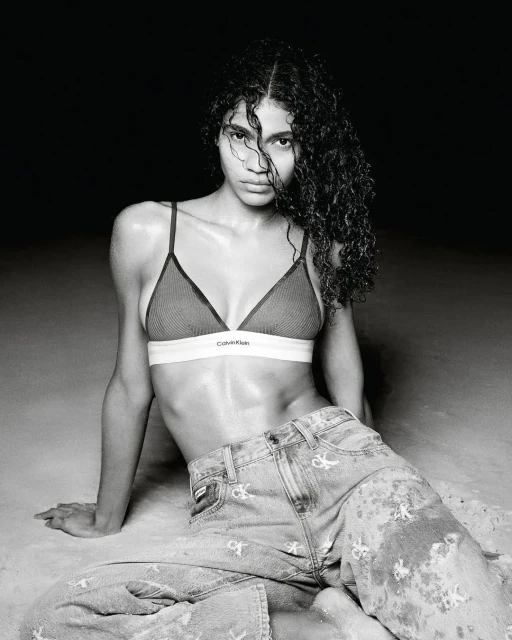 Model with dark curly hair wearing a Calvin Klein bralette and monogram denim jeans seated on a dark ground.