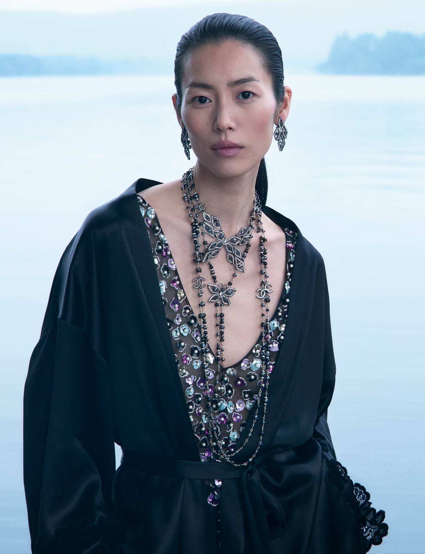 Model Liu Wen wearing a black silk robe and elaborate layered necklaces against an outdoor water background for Chanel