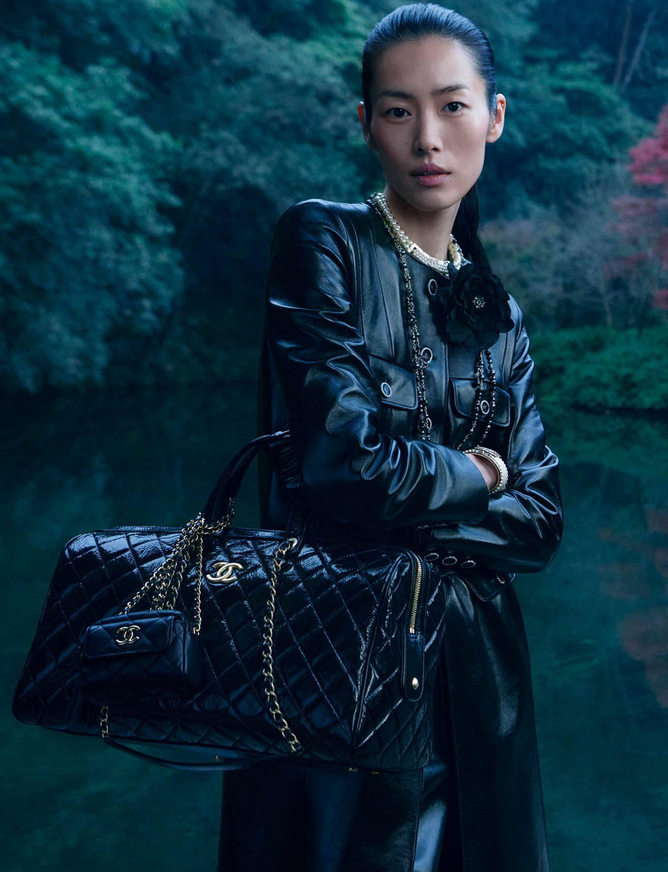 Model Liu Wen holding a black quilted leather handbag outdoors by water for the Chanel 2024/25 Metiers d'Art campaign.