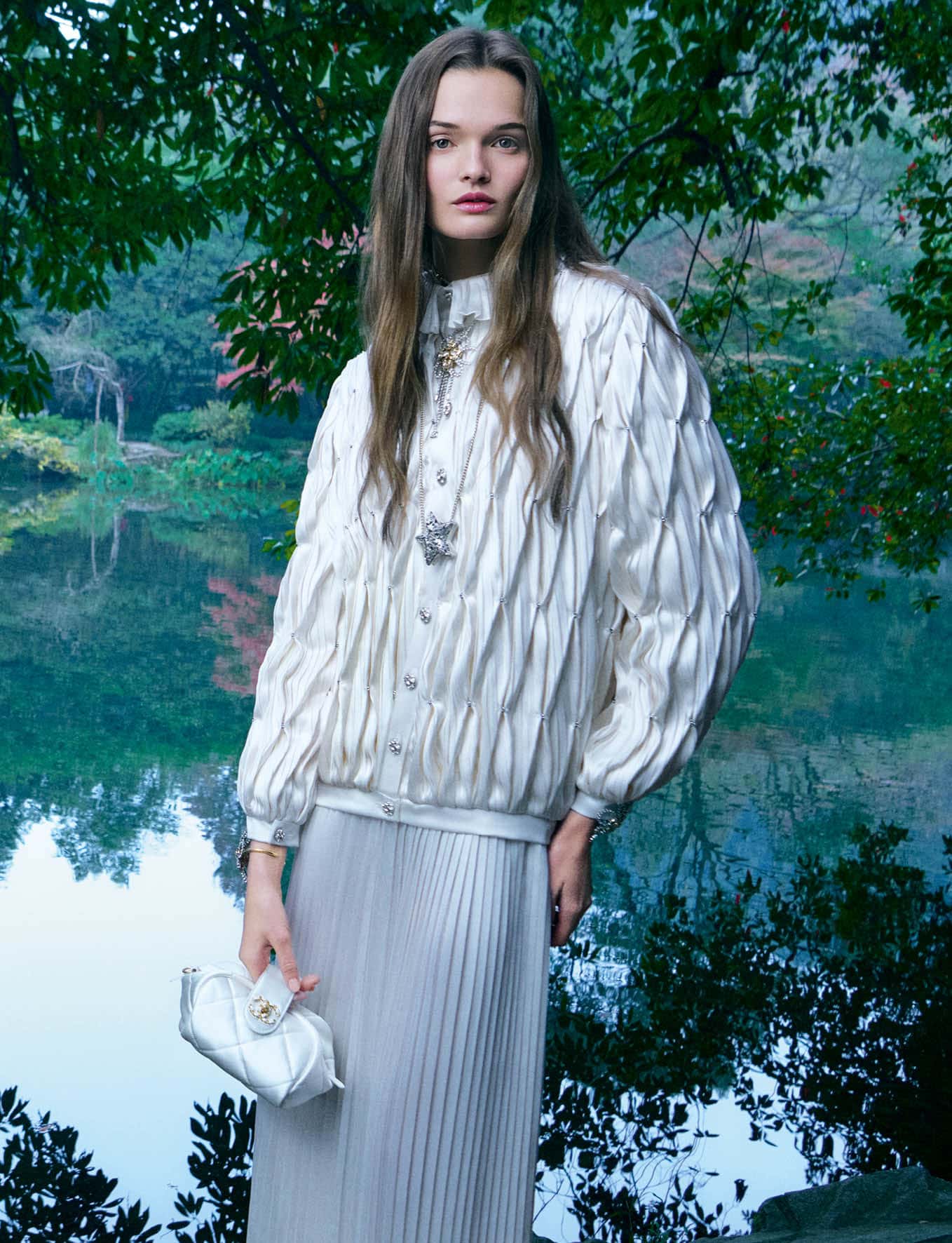 Model wearing a white textured jacket and pleated skirt holding a white handbag outdoors near a lake