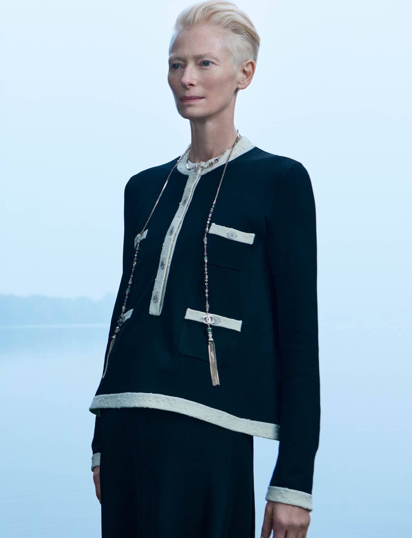 Tilda Swinton wearing a dark cardigan with white trim against a hazy light blue background for Chanel