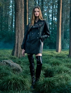 Model standing in a dense forest wearing a dark textured coat and tall patent leather boots for Chanel campaign
