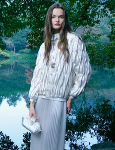 Model wearing a white textured jacket and pleated skirt holding a white handbag outdoors near a lake