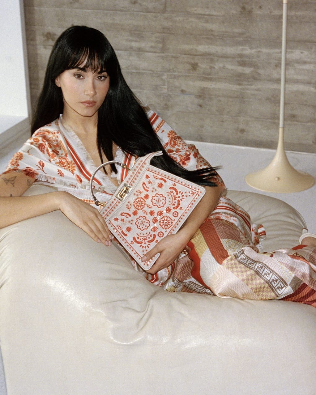 Model with long dark hair holding a patterned handbag while seated on a white leather pouf against a concrete wall
