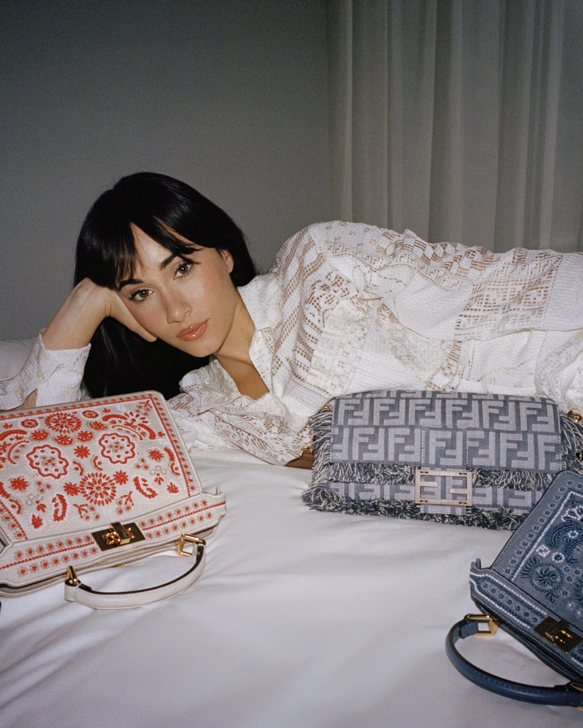 Model wearing white lace shirt reclining on white bedding with embroidered Fendi handbags in a bedroom